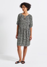 Schwarz-weißer gemusterter Kurzarmkleid mit einem Schlüssellochausschnitt, gerafftem Bund und lockerer Passform, kombiniert mit schwarzen Slipper-Sandalen.