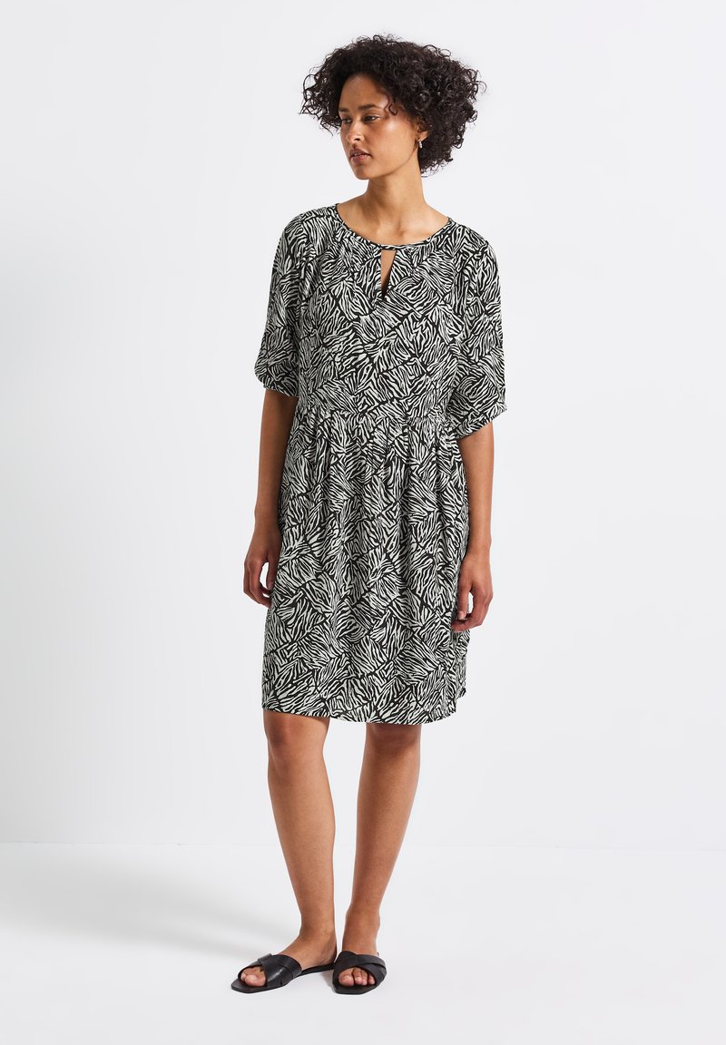 Schwarz-weißer gemusterter Kurzarmkleid mit einem Schlüssellochausschnitt, gerafftem Bund und lockerer Passform, kombiniert mit schwarzen Slipper-Sandalen.