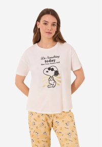 Witte katoenen t-shirt met korte mouwen, met zwart tekst en een Snoopy-illustratie met gouden accenten. Gekleed met gele broeken met een patroon.