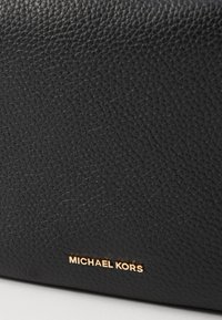 Černá texturovaná kožená taška s pozlaceným vyraženým logem "MICHAEL KORS". Povrch ukazuje jemný vzor zrnění.