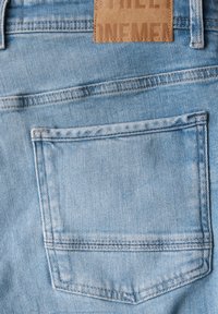 Vaaleansininen farkkukangas, jossa on takatasku vahvistetulla reunalla ja sen ylle ommeltu nahkainen etiketti, jossa lukee "STREET ONE" brändäystä varten.