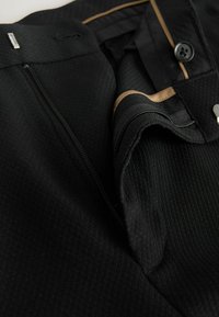 Czarna odzież z teksturowanej tkaniny, z zamkiem błyskawicznym i detalami na guzik, z złotymi akcentami wzdłuż wewnętrznej szwy. Zbliżenie na wzór.