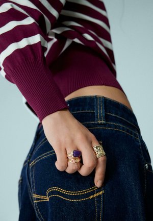 Sötétkék farmert és bordó csíkos pulóvert viselő személy, keze a hátsó zsebén pihen, ahol három különálló aranygyűrű látható.