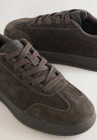 Ciemnobrązowe zamszowe sneakersy z dopasowanymi sznurówkami i czarnymi gumowymi podeszwami, pokazane z bliska pod kątem na jasnym tle.