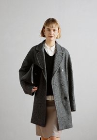 Palton gri din lână, cu un design cu două rânduri de nasturi, rever în formă de U și două buzunare frontale. Asociat cu un pulover negru și o fustă bej.