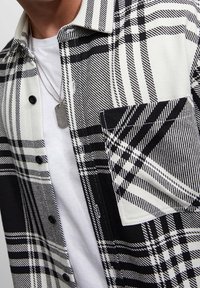Camicia a quadri bianco e nero realizzata in tessuto con chiusura a bottone, una singola tasca sul petto e una texture liscia. Indossata sopra una camicia bianca.