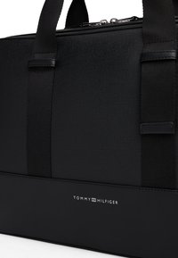 Black duffel bag made of textured fabric and smooth leather, featuring dual handles and a silver logo. Zipper closure at the top.