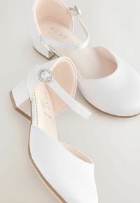 Valkoiset avonaiset kengät matalahkolla korolla, säädettävällä nilkkahihnalla ja koristeellisella kimallekivellä. Silkkinen pinta ja yksinkertainen muotoilu.