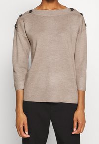 Beige pletený svetr s širokým výstřihem, tříčtvrtečními rukávy a velkými černými knoflíky na ramenech. Texturovaná látka s uvolněným střihem.