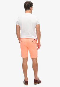 T-shirt bianco da uomo abbinato a pantaloni corti arancioni brillanti. I pantaloni hanno due tasche posteriori con bottoni neri e un taglio dritto.