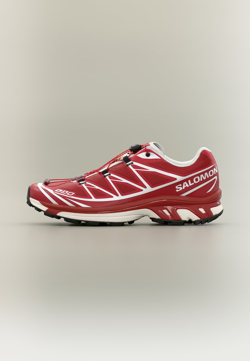 Zapatilla de trail running Salomon en rojo y blanco con suela negra, sistema de cordones rápidos y patrones geométricos laterales sobre un fondo neutro.