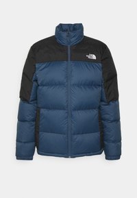 Veste matelassée bleu et noire avec texture matelassée, col montant, fermeture éclair intégrale et logo sur la poitrine. Dotée de panneaux latéraux noirs.
