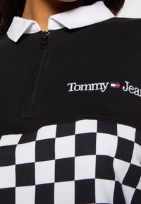 Haut à fermeture éclair à carreaux noir et blanc avec un col blanc, portant "Tommy Jeans" brodé en fil blanc sur la poitrine.