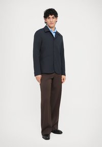 Tengerészkék, gombos dzseki hegyes gallérral, lapos első zsebekkel, barna nadrággal és fekete cipővel párosítva. Simább, strukturált anyag.