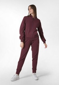 Bordeauxkleurige zip-up jas en bijpassende jogger set, gemaakt van zachte stof met ribbels op de manchetten, met een merkaccent op de jas. Witte sneakers.