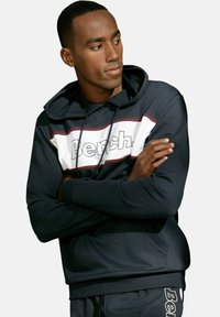 Marineblaue Kapuzenjacke mit einem weißen und kastanienbraunen Streifenmuster, das das Wort "Bench" in fetten Buchstaben enthält. Weiches Material, verstellbare Kapuze mit Kordelzug.