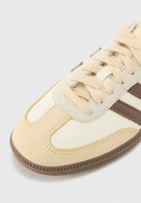 Baskets beige et crème avec des rayures marron, bout rond, semelle en caoutchouc texturée et système de laçage. Matériaux doux avec des accents cousus.