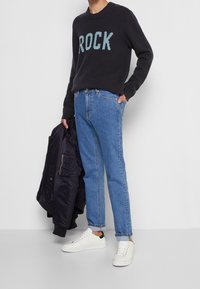 Dunkel gestrickter Pullover mit "ROCK" in hellblauen Buchstaben, kombiniert mit hellblauen Jeans und weißen Sneakers. Hält eine dunkle Bomberjacke.