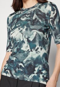 Stickad topp med kort ärm i ett abstract mönster i teal och svart. Slät textur med en figurnära design och rundad hals.