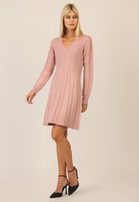 Hellrosa gestricktes Kleid mit V-Ausschnitt und langen Ärmeln, das ein Zopfmuster und einen ausgestellten Saum aufweist. Kombiniert mit schwarzen High Heels.