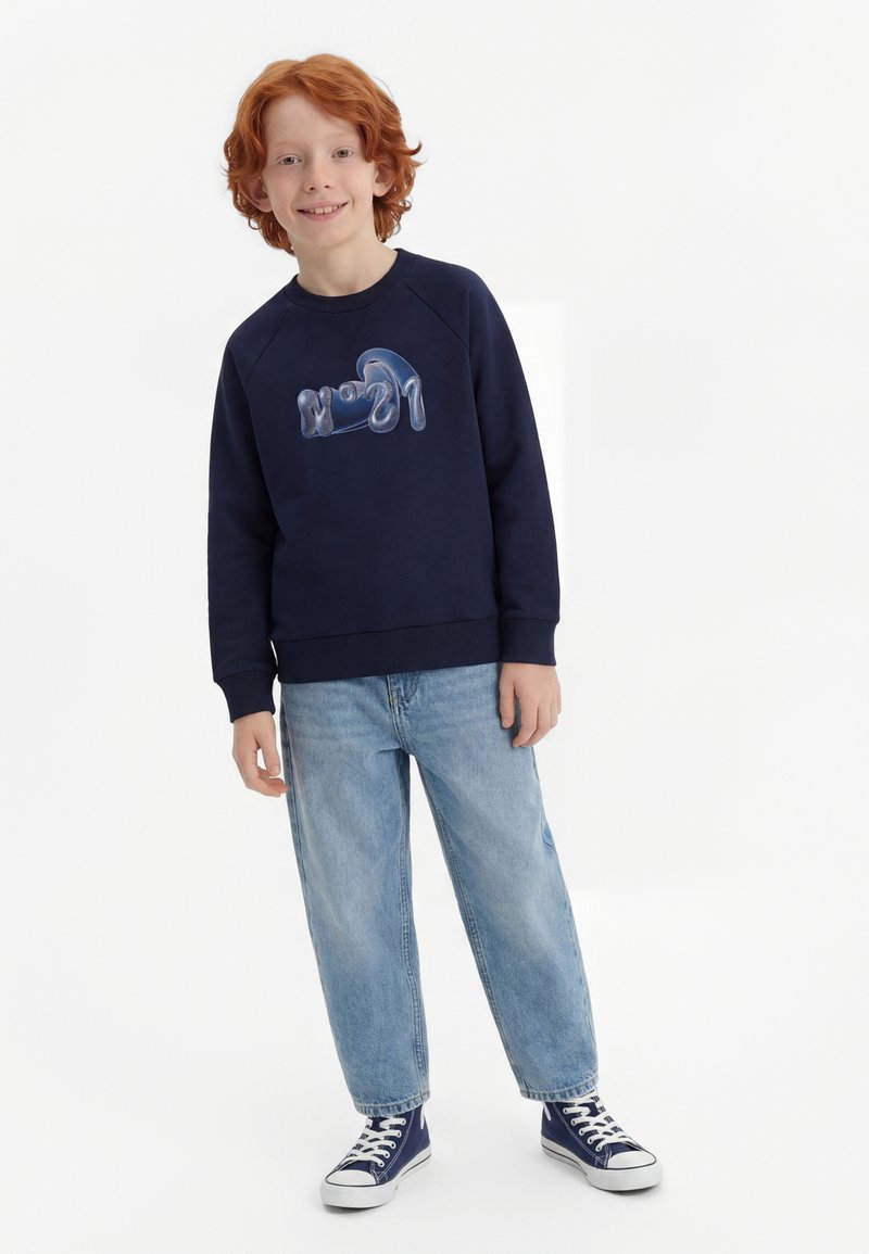 Rothaariges Kind lächelnd, trägt einen marineblauen Pullover mit blauem "N°21"-Schriftzug, hellblaue Jeans und marineblaue High-Top-Sneaker auf weißem Hintergrund.