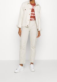 Veste en denim blanc, t-shirt rouge tie-dye, pantalon beige clair et baskets blanches. La veste possède de grandes poches sur la poitrine et des boutons en cuivre.