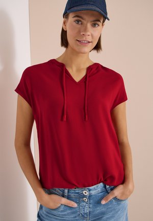Vrouw met een blauwe baseballpet, een rood kort mouwloos capuchonshirt en lichtblauwe jeans, staand met de handen in de zakken tegen een lichte achtergrond.