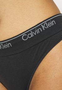Musta puuvillabikiinialusvaate, jonka pinnassa on sileä rakenne ja joustava vyötärö, jossa on harmaa "Calvin Klein" -logo. Lähikuva, joka korostaa ompelutietoja.