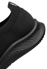 Detailní záběr paty tenisky z černé pletené látky s žebrovaným límcem kolem kotníku a podešví s texturovaným vzorem.