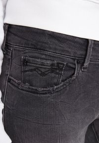 Černé denimové džíny se zničenou zadní kapsou, viditelným šitím a mírným vyblednutím. Pas je spojen s bílým vnějším oděvem.