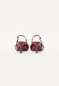 Escarpins en cuir verni rouge avec un thin stiletto, un dos ouvert et un petit nœud sur les côtés. Texture lisse et brillante.