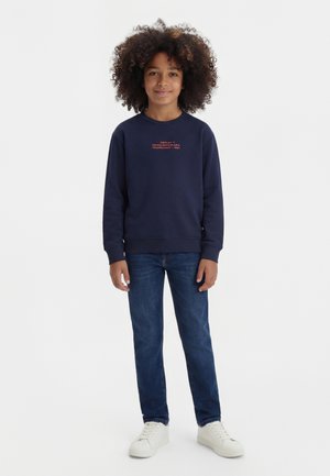 Ragazzo con capelli ricci che indossa una felpa blu navy, jeans blu e scarpe da ginnastica bianche, in piedi e sorridente con uno sfondo bianco semplice.