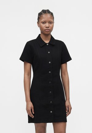 Femme aux cheveux tressés portant une robe courte noire boutonnée avec manches courtes, debout devant un fond clair uni.
