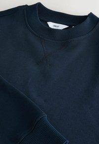 Sweat-shirt bleu marine à col rond avec col et poignets côtelés, avec un détail X cousu sous l'encolure et une étiquette de marque blanche à l'intérieur du col.