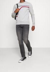 Szary sweatshirt z czarnymi, czerwonymi i białymi paskami na klatce piersiowej, w zestawieniu z ciemnoszarymi dżinsami i czarnymi sneakersami, model w ruchu.