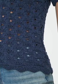 Jūras spēku zils adīts tops ar teksturētu atvērtu rakstu, ar viļņotu apakšmalu un īsām piedurknēm, kombinēts ar džinsu biksēm.