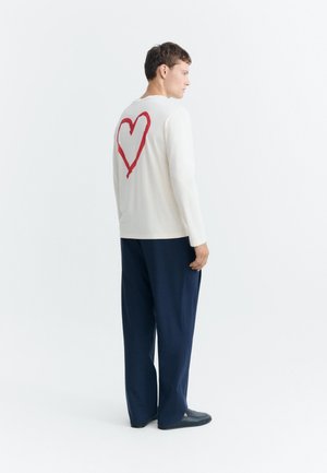 Homme portant une chemise blanche à manches longues avec un grand cœur rouge dans le dos, pantalon bleu marine et chaussures noires, debout devant un fond blanc.