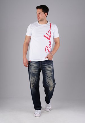 Mann in einem weißen T-Shirt mit roten vertikalen Text und ausgebleichten dunkelblauen Jeans, der vor einem grauen Hintergrund steht und nach links schaut.