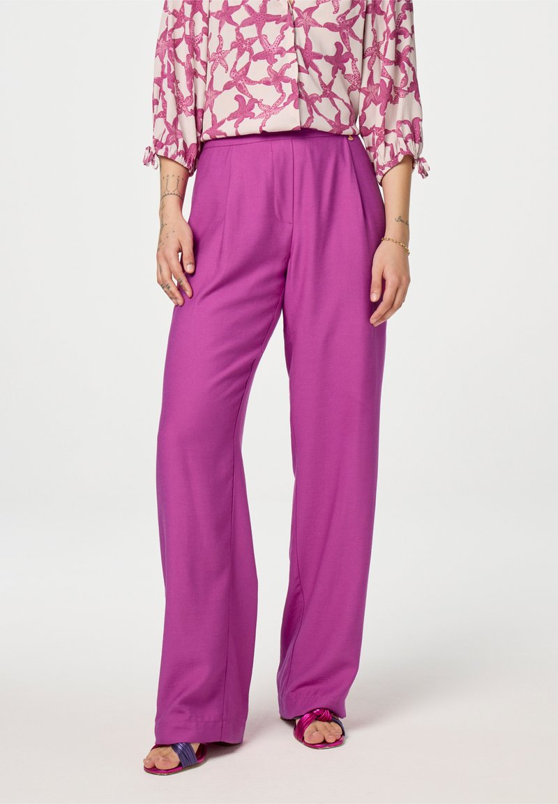 Vrouw in hoge taille, wijde magenta broek, gebloemd roze blouse met geplooide mouwen, paarse en roze open-toe sandalen en polsjuwelen.