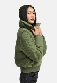 Olive green zip-up jacket with a plush, fleece-like body and quilted sleeves. Features a high collar and ribbed hem.
