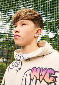 Beżowy hoodie z czarnym nadrukiem uśmiechniętej buźki, z kolorową grafiką "NYC". Gładka tekstura, duża kaptura i ściągacz przy kołnierzu.