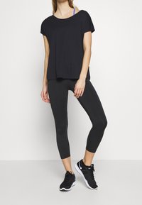 Mujer con leggins negros cortos, camiseta negra holgada, zapatillas negras y una correa de sujetador deportivo azul visible, de pie contra un fondo blanco.