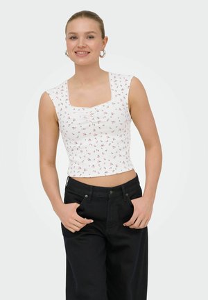 Young woman smiling, wearing a white floral sleeveless crop top and black high-waisted pants, hands in pockets against a plain background.