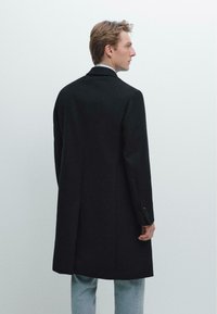 Mann in einem schwarzen langen Mantel und hellblauen Jeans, der seitlich vor einem schlichten weißen Hintergrund steht.