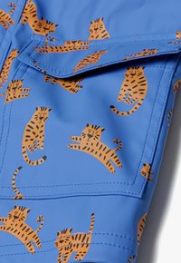 Blåt vandtæt stof med et gennemgående mønster af orange tigre med sorte striber. Synlig lomme med syninger som detaljer.