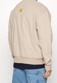 Beige sweatshirt met geribbelde boorden en onderrand, heeft een geel label op de achterkant van de halslijn. Zachte stof, comfortabele pasvorm en eenvoudig ontwerp.