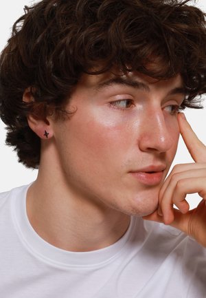 Jeune homme avec des cheveux bouclés et foncés, portant un t-shirt blanc, arborant une petite boucle d'oreille noire en forme d'étoile. Vue de profil en gros plan, fond neutre.