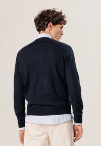 Pull marine en tissu doux, doté de manches longues et d'un col rond. Superposé sur une chemise à rayures bleues et blanches.