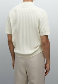 Homme portant un polo à col en tricot côtelé à manches courtes de couleur écrue et un pantalon beige, vu de dos sur un fond clair.