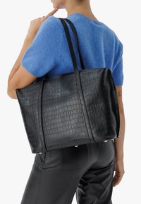 Sac fourre-tout noir en cuir avec embossage crocodile et poignées doubles. Fabriqué en cuir, avec une surface texturée. Porté sur un haut en tricot bleu.